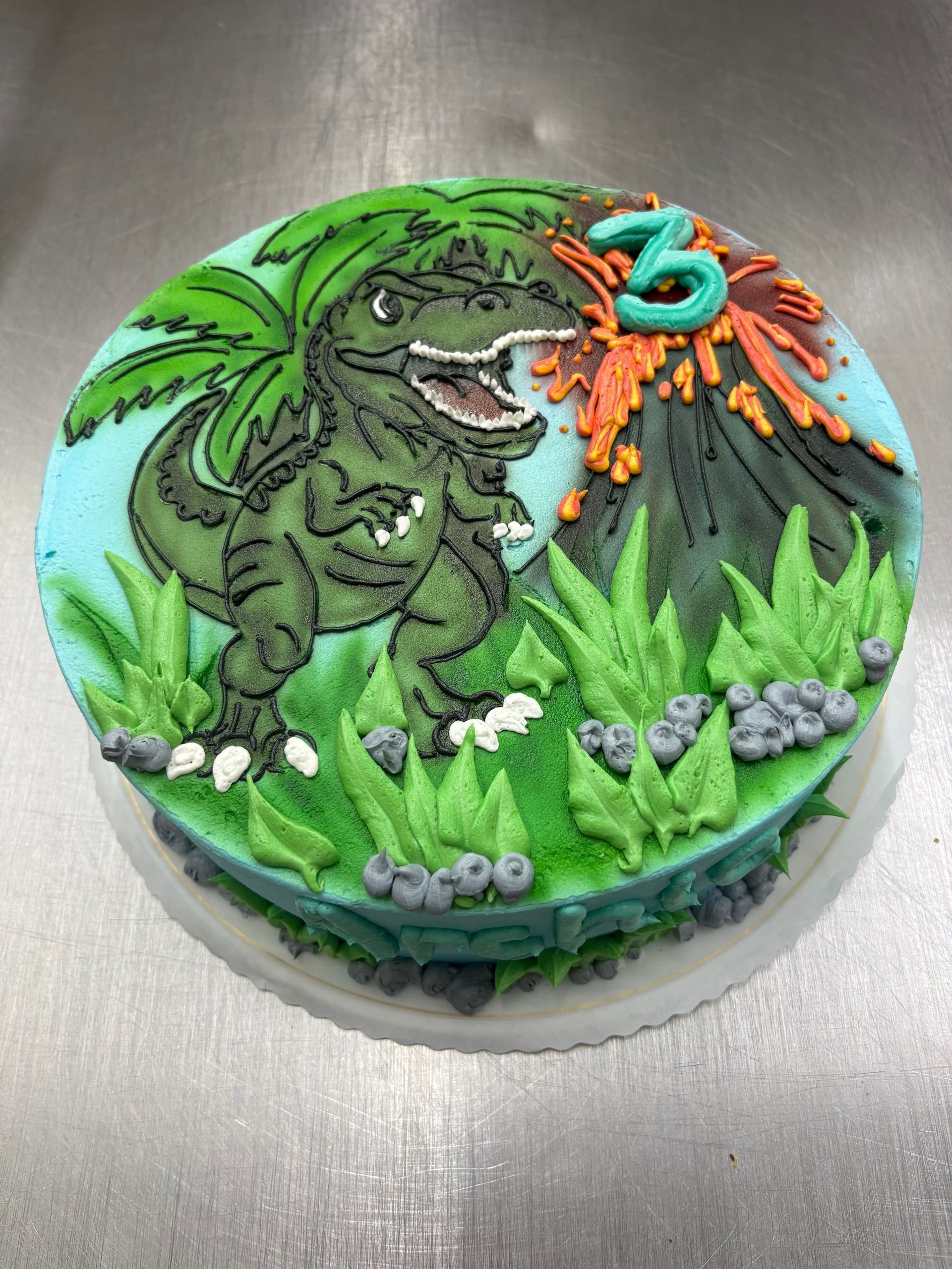 Dinosaur-themed birthday cake with green T-Rex, volcano, number 3, and green and blue frosting.