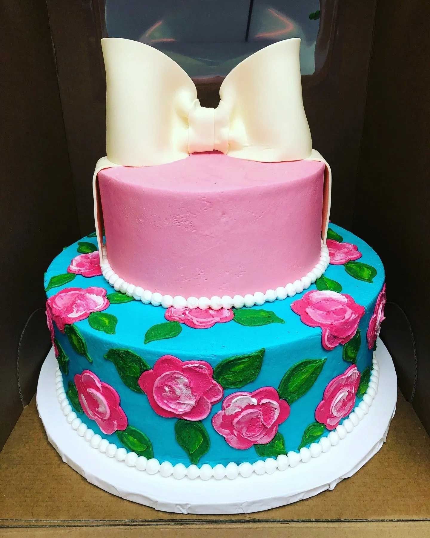 Two-tiered cake with pink frosting on top tier and turquoise with pink roses on bottom. A large white bow sits atop.