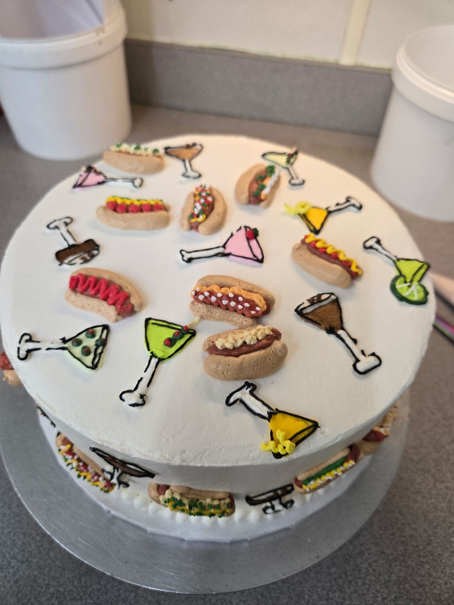 Two-tiered cake decorated with hot dogs and cocktail-themed icing designs on a white background.