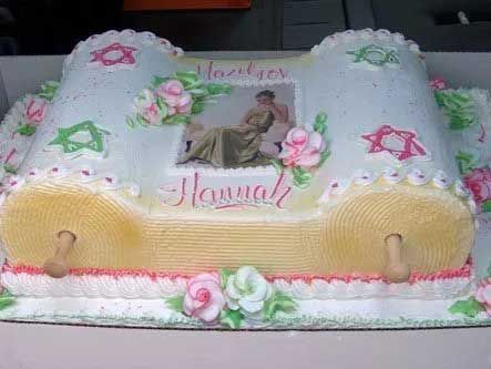 Cake shaped like a Torah scroll, decorated with frosting, a photo, and floral accents.