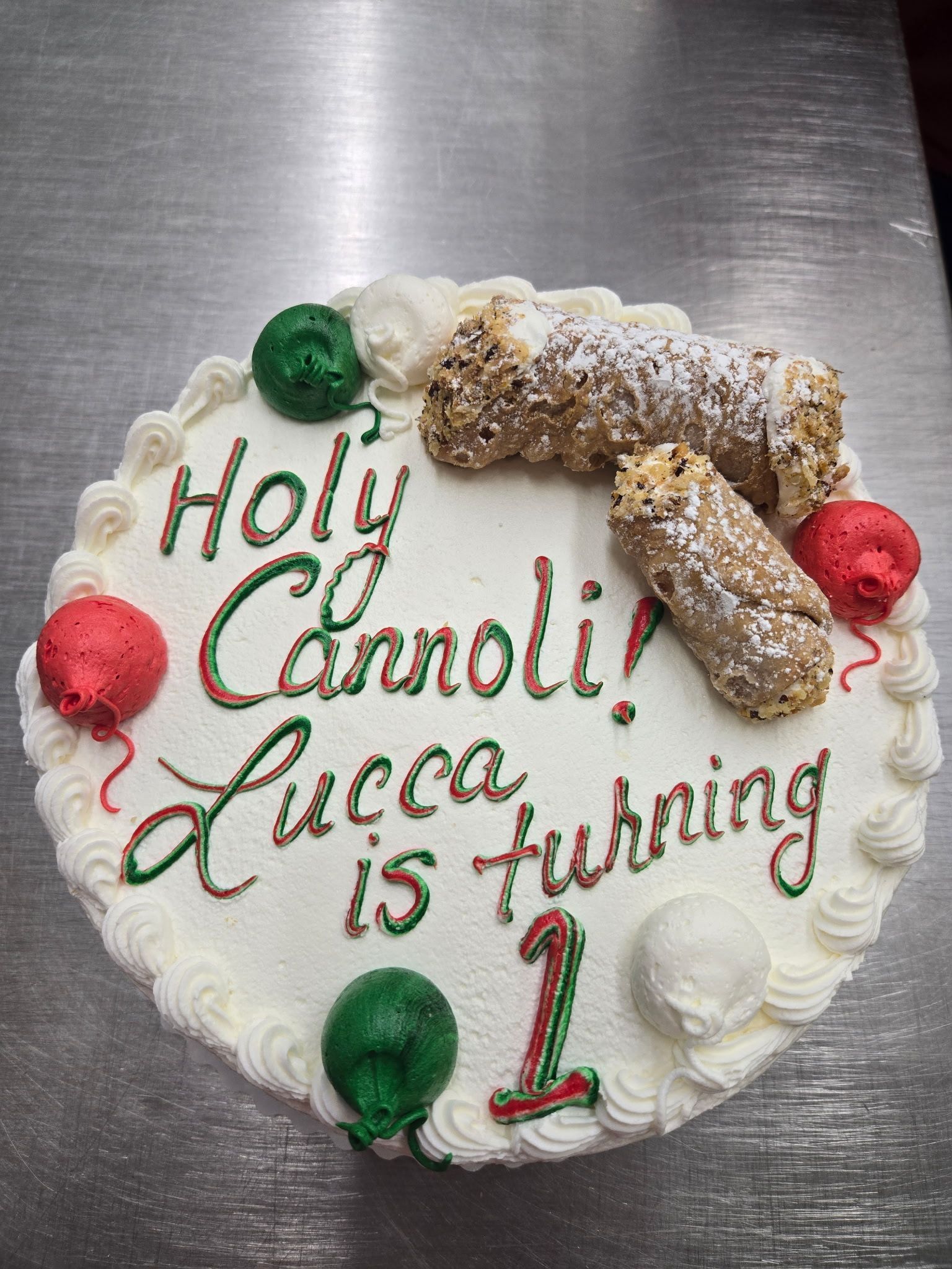 Cake with Holy Cannoli! Lucca is turning 1 in red and green, topped with cannoli and balloons.