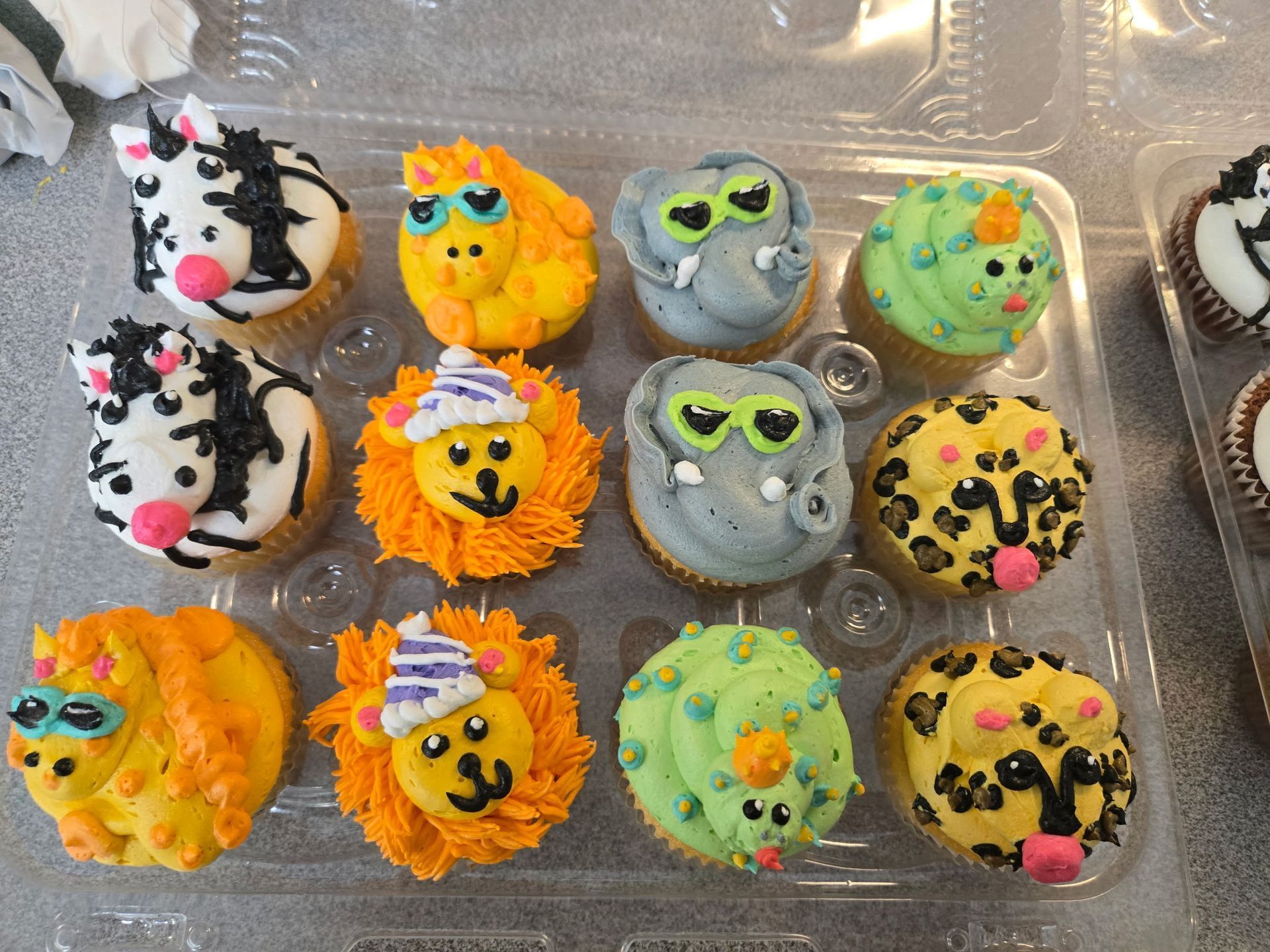 Animal-themed cupcakes in a plastic container; zebra, lion, elephant, unicorn, and leopard decorated with frosting.