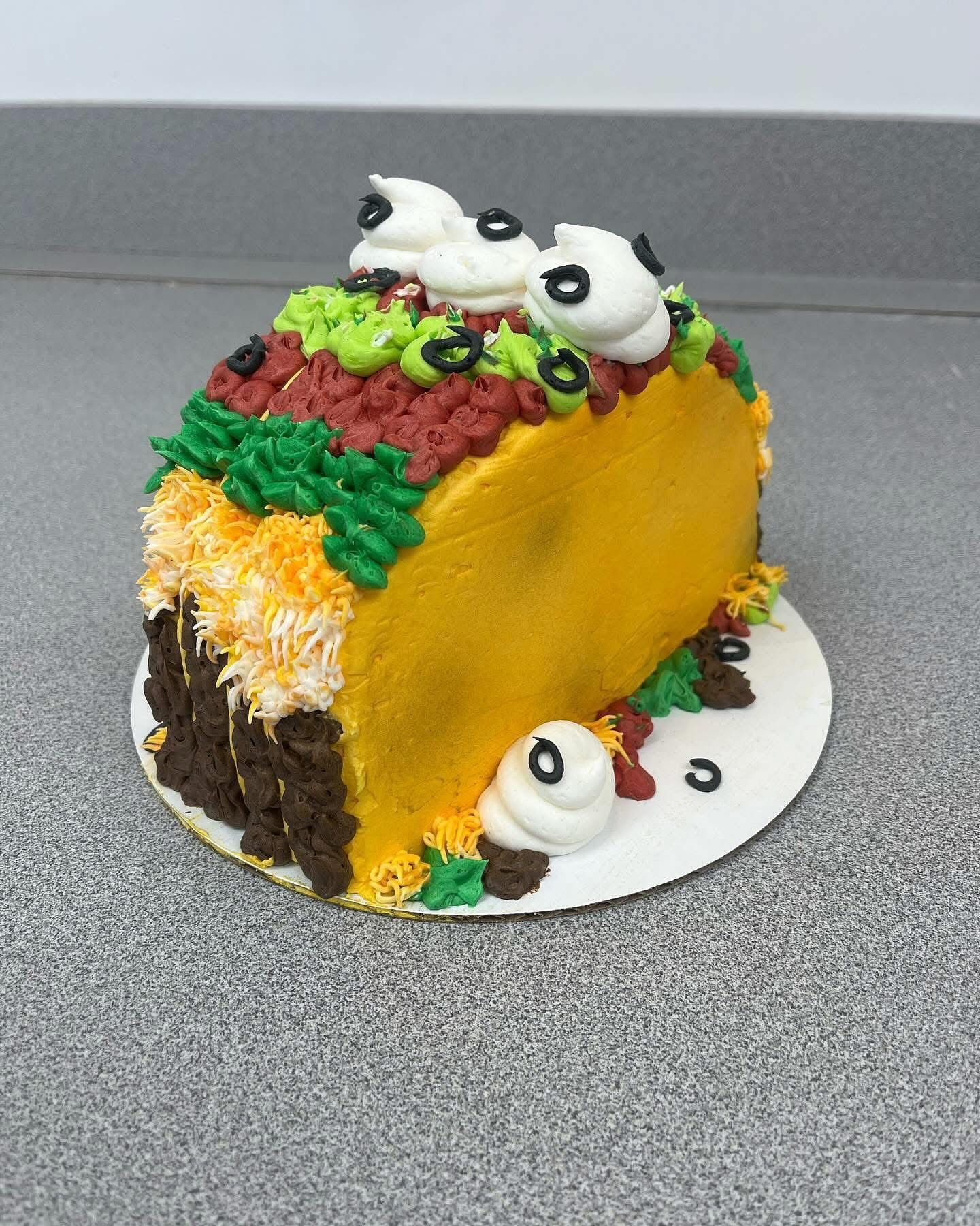Taco-shaped cake with buttercream toppings like lettuce, cheese, meat, olives, and sour cream, on a white plate.