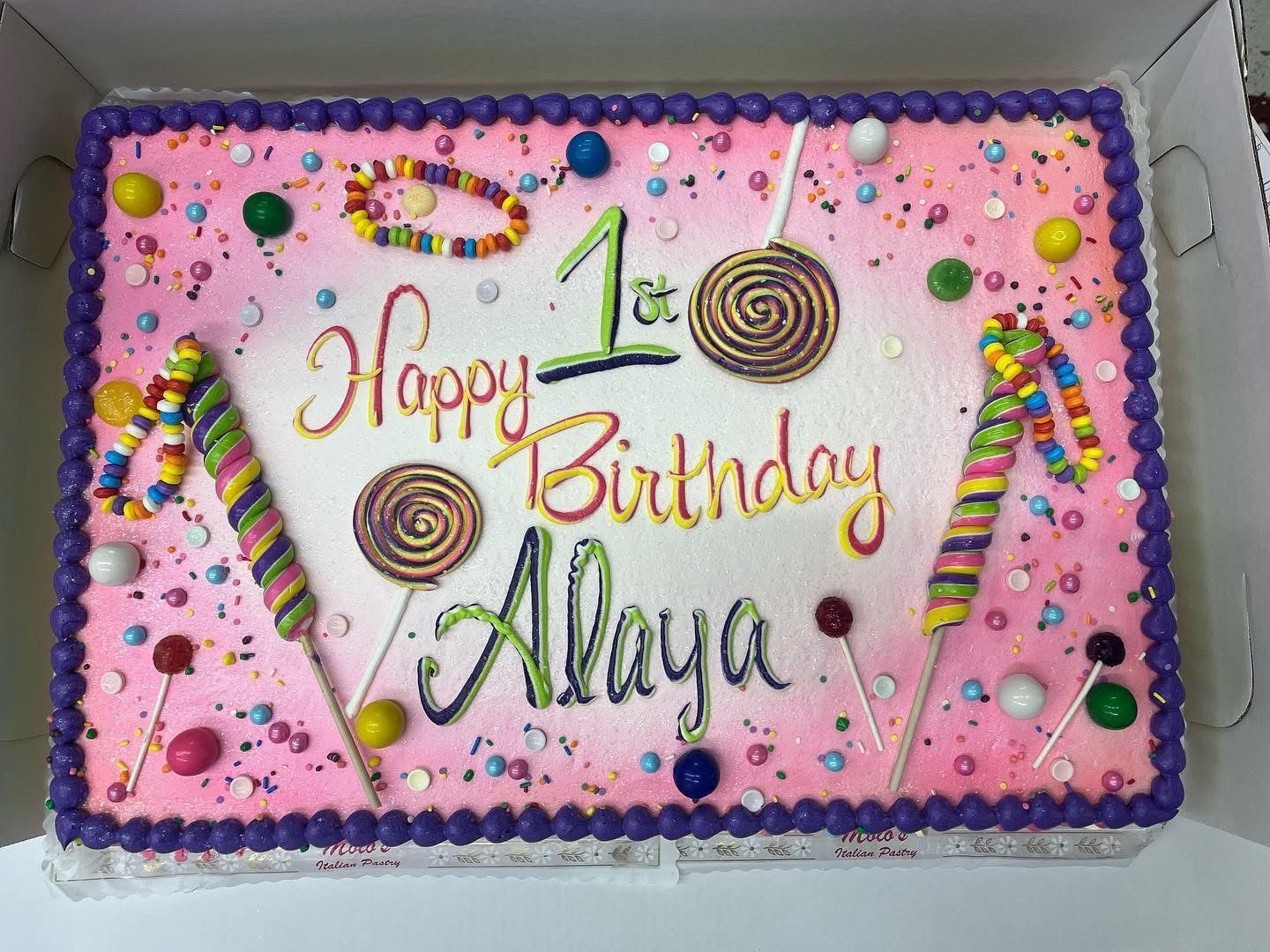 Birthday cake for Alaya, decorated with lollipops, candy, and colorful sprinkles, celebrating a first birthday.