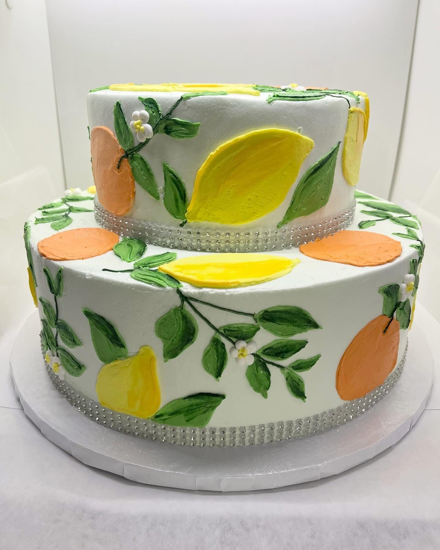 Two-tiered white cake decorated with painted lemons, oranges, and green leaves, and bedazzled edges.