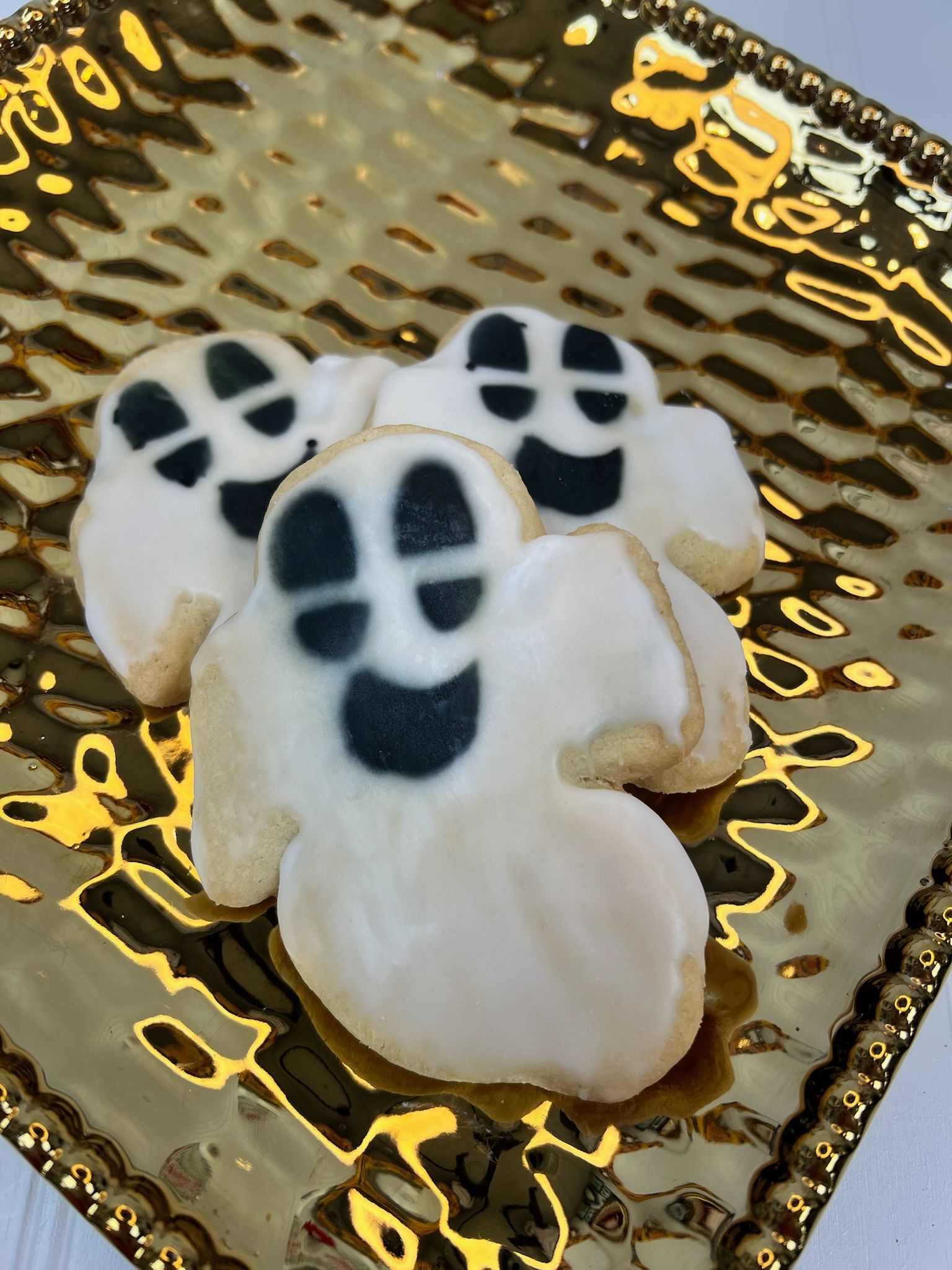Three ghost-shaped cookies decorated with white icing and black facial features on a gold, textured plate.