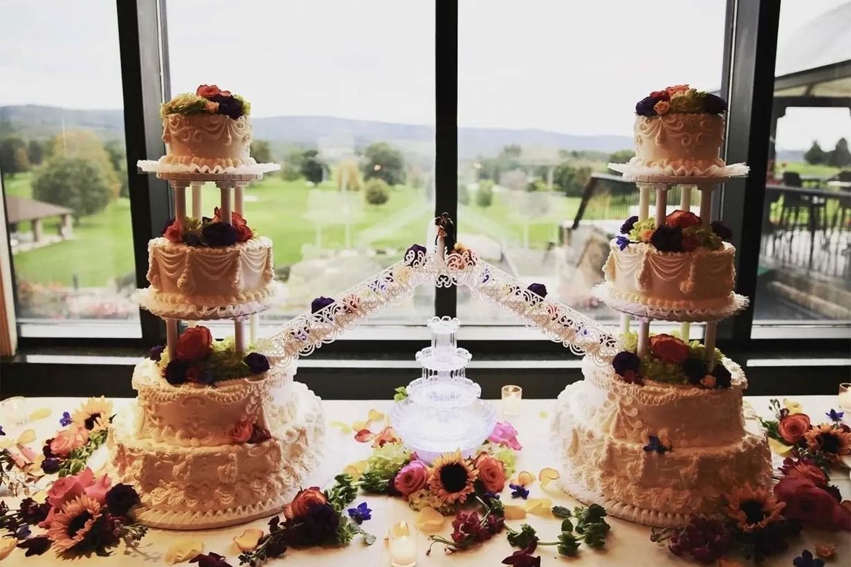 Two ornate tiered wedding cakes with fruit atop, connected by a decorative structure, set before a window view.