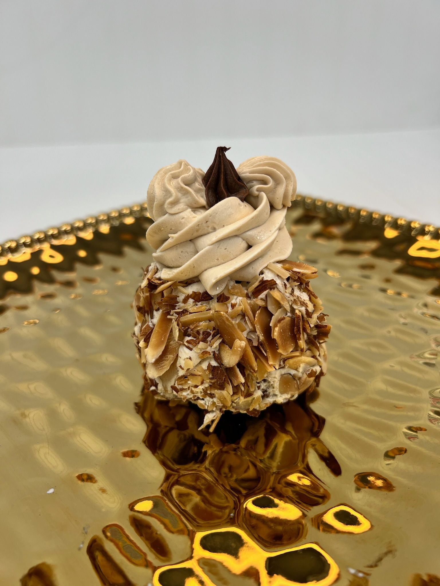 Dessert on a gold plate: Round cake with almond flakes, whipped cream topping, and chocolate garnish.