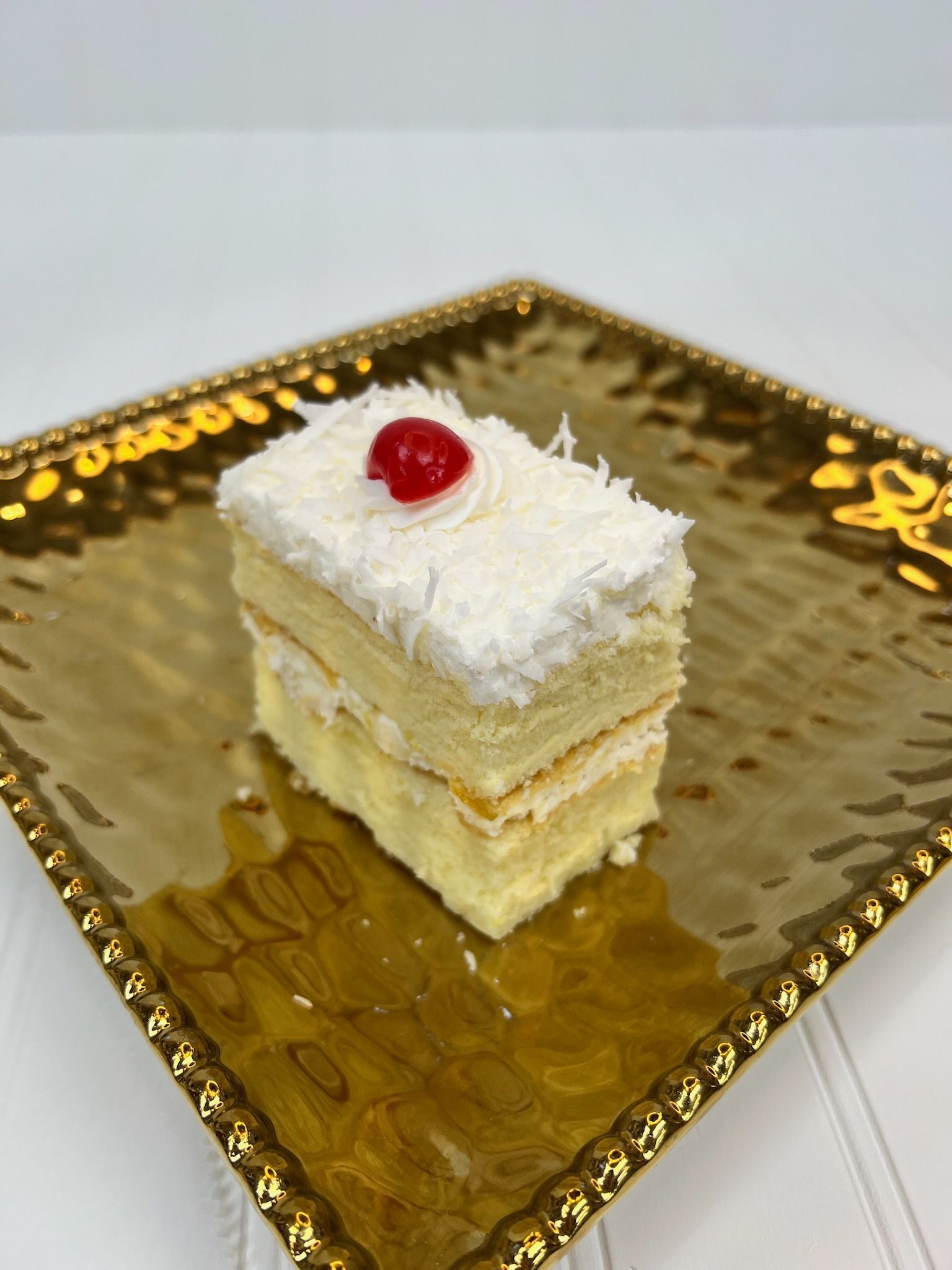 Slice of layered cake with shredded coconut and a cherry on a gold, textured plate.