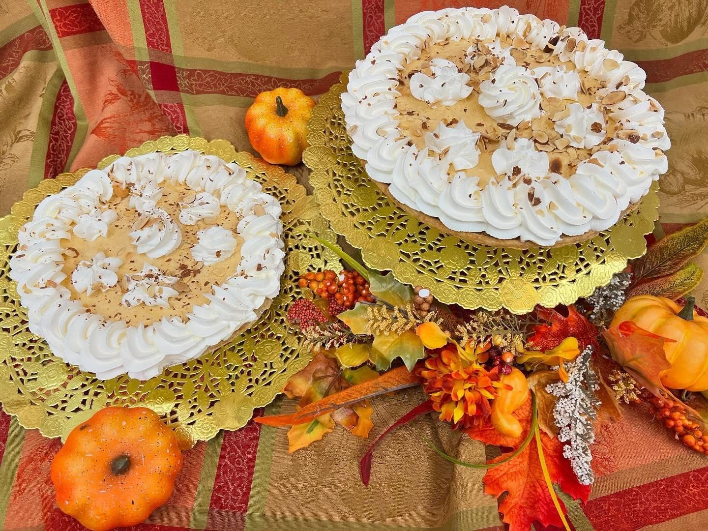 Two pumpkin pies with whipped cream on decorative golden doilies, surrounded by fall foliage and pumpkins.