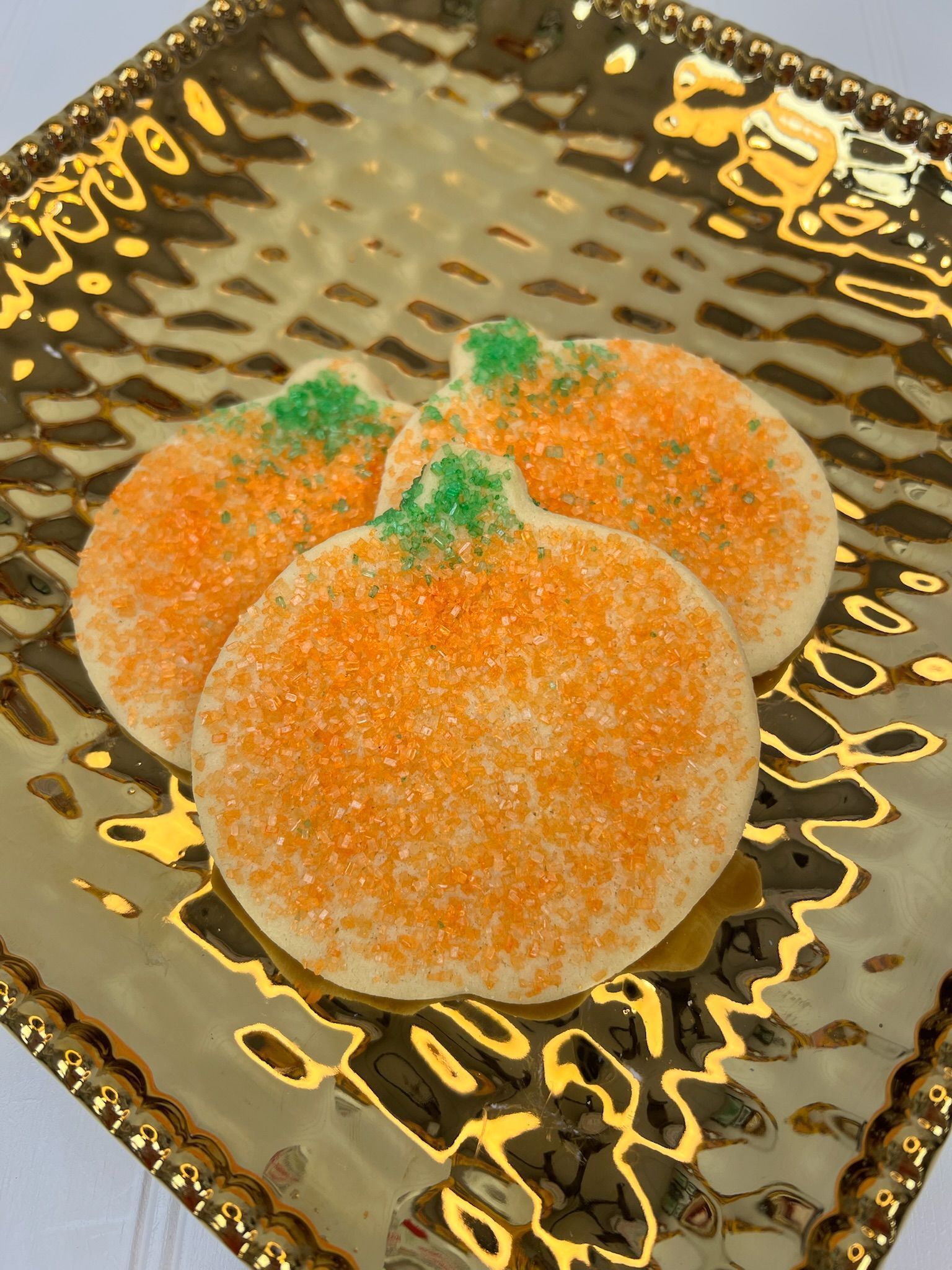 Pumpkin-shaped cookies with orange sugar coating and green sprinkle stems, arranged on a gold, textured plate.