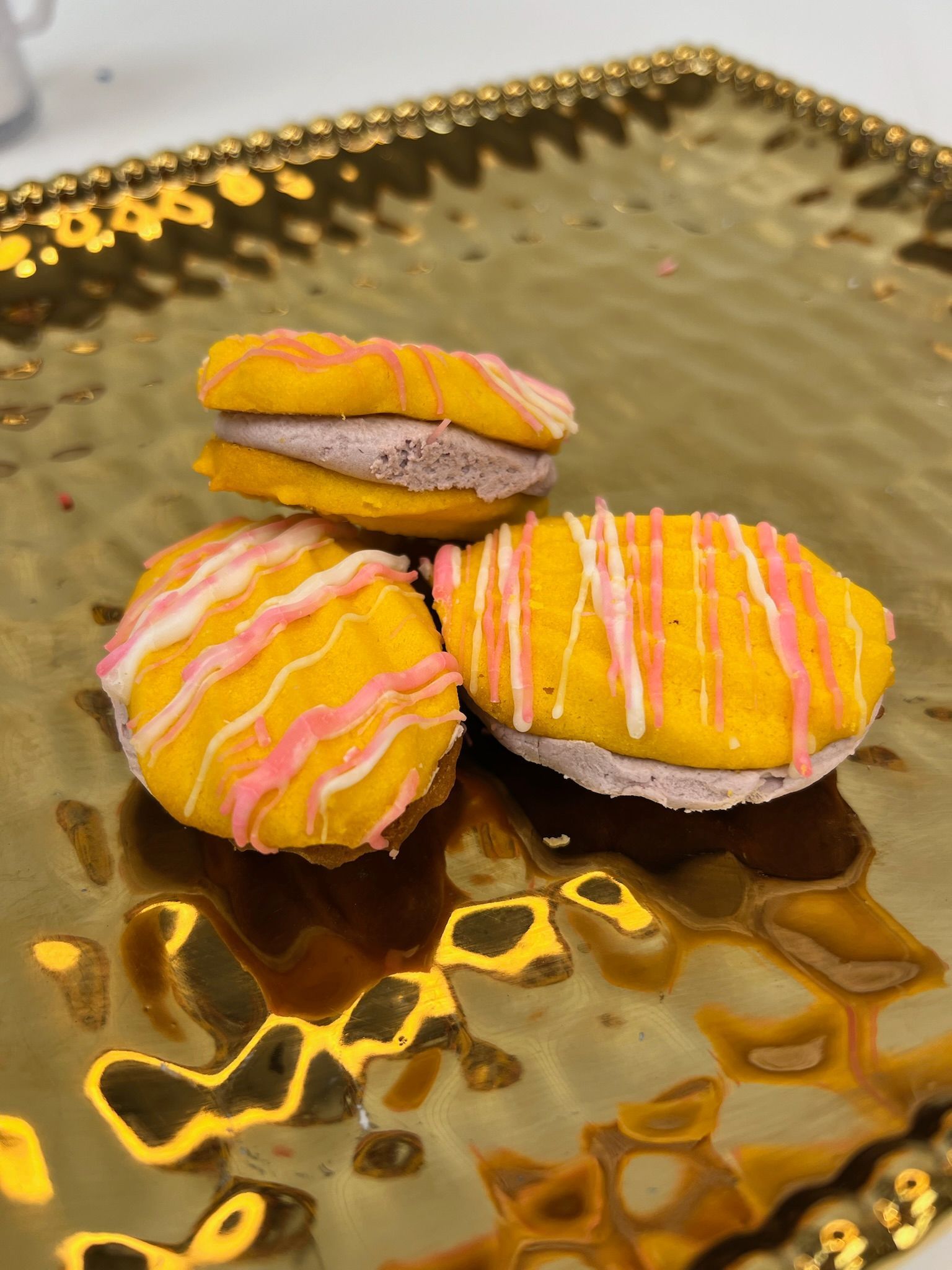 Three colorful macarons on a gold tray. Yellow shells with pink and white drizzled icing, filled with lavender cream.