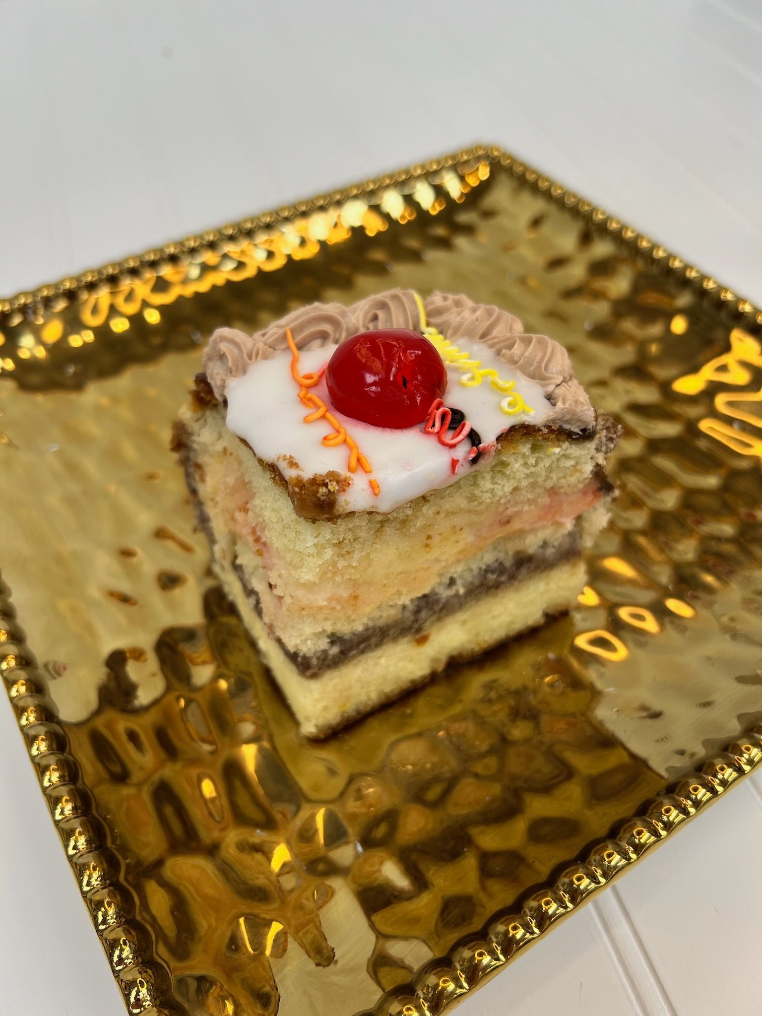 Slice of layered cake on gold plate, topped with cherry and frosting.