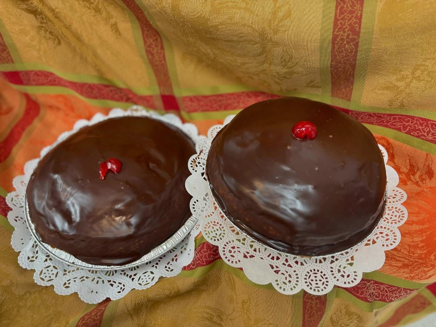 Two chocolate-covered cakes on decorative paper doilies, each topped with a cherry, on a colorful plaid surface.