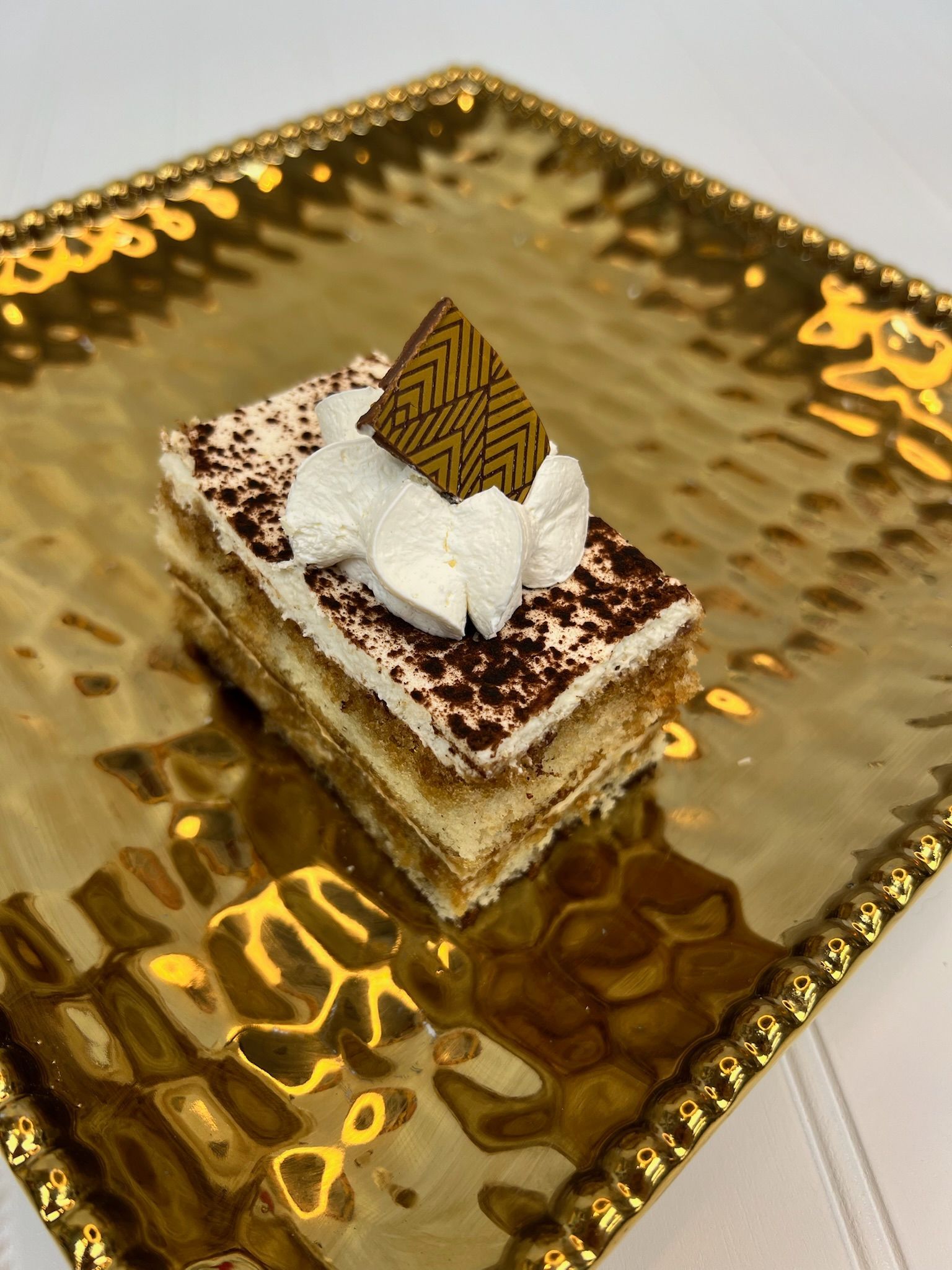Tiramisu dessert on a gold decorative tray, topped with whipped cream and chocolate garnish.