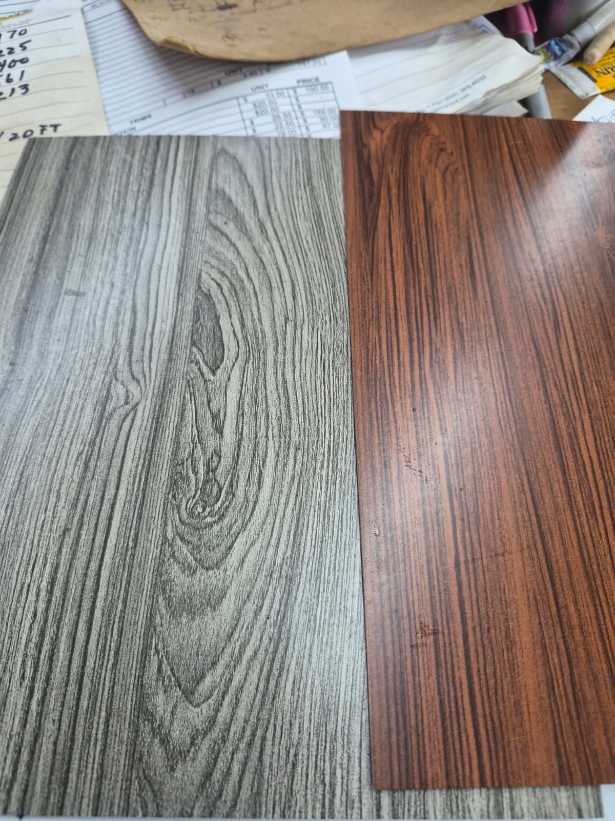 Two wood-grain flooring samples, one gray and one reddish brown, laid side by side on a tabletop.