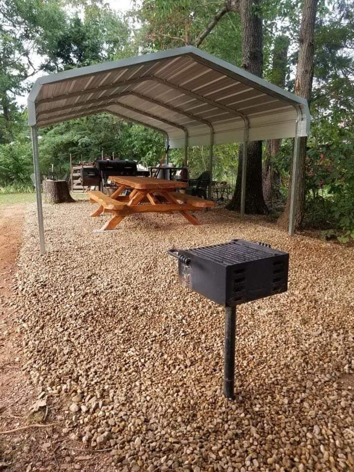 Covered picnic area with wooden tables, gravel ground, and a black grill in a wooded park.