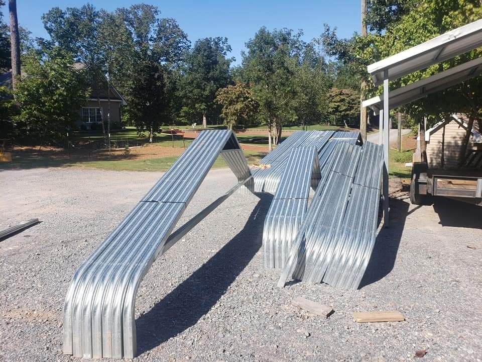 Two curved metal ramps on gravel in a sunny outdoor yard, with trees and a covered structure nearby