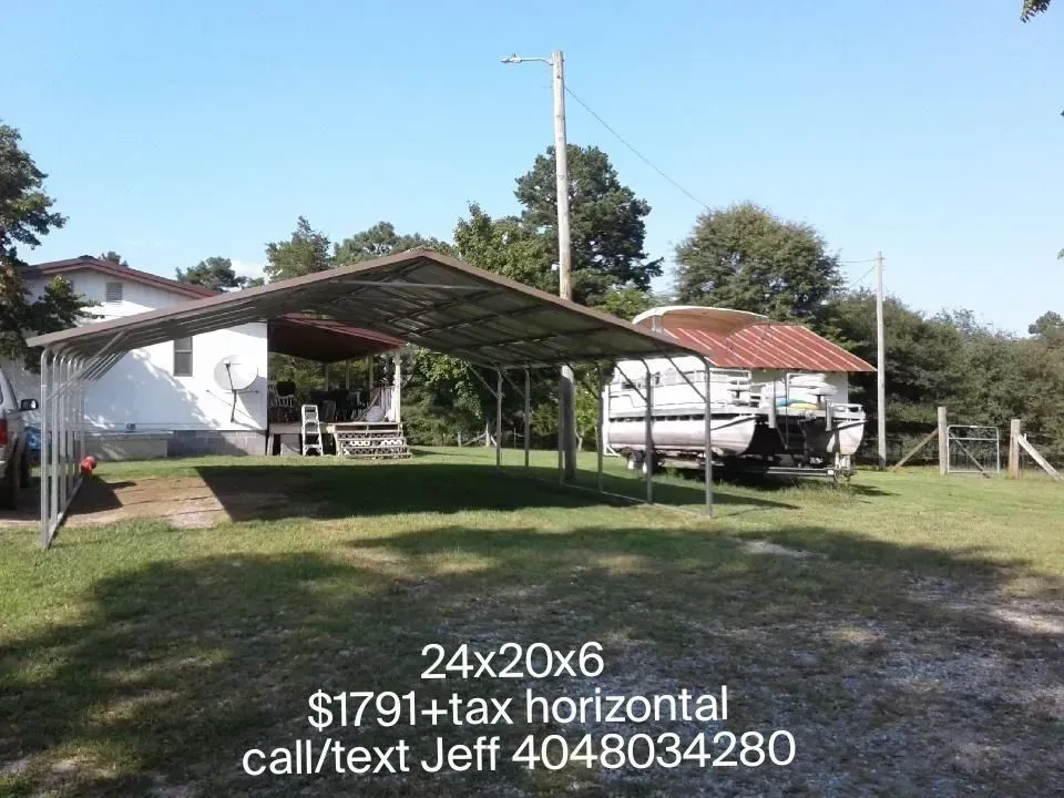 Grassy yard with a metal carport, parked trailer, and small white building under a clear blue sky