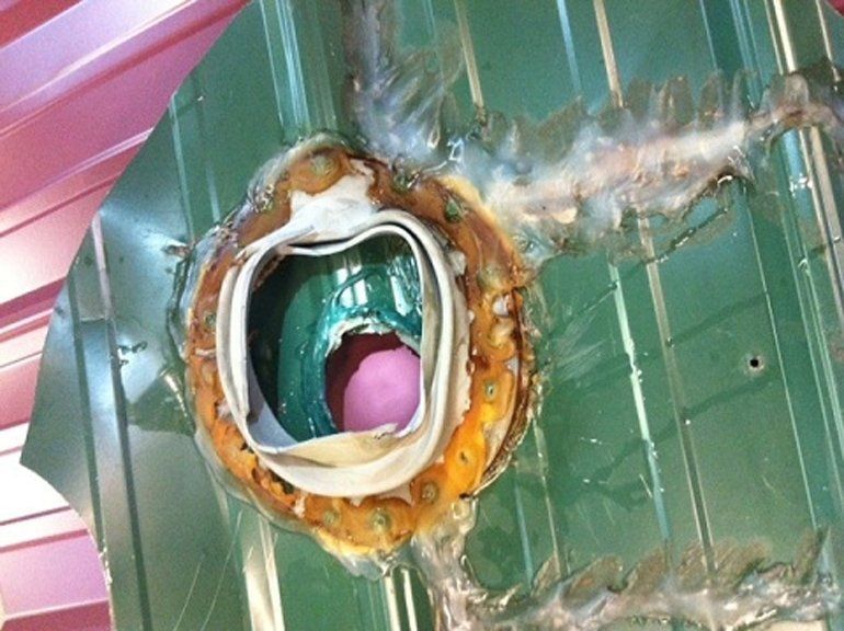 Severely damaged green metal door with a circular hole and torn, rusted edges, exposing a pink object inside