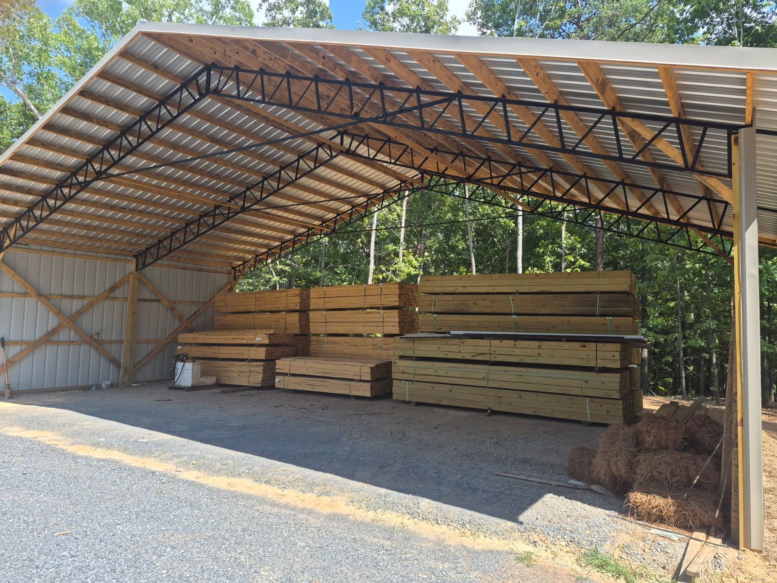 Covered lumber storage shed with stacked wooden beams on gravel beside trees