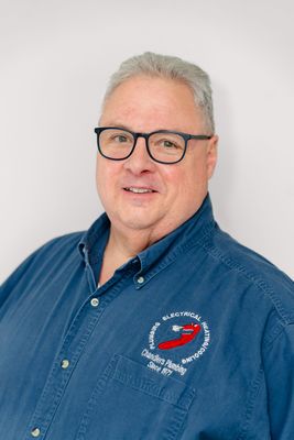 A person wearing glasses and a blue button-down shirt with an embroidered logo on the chest, smiling against a white wall.