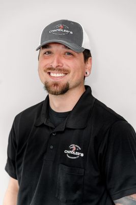 A smiling person with a goatee wearing a gray cap and black polo shirt with a logo, standing against a white background.