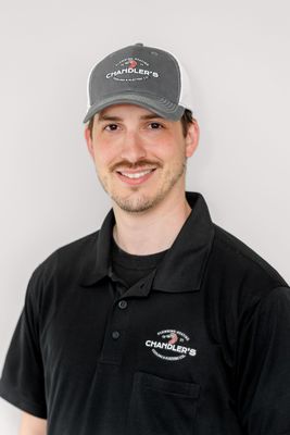 A person smiling while wearing a grey baseball cap and a black polo shirt, both featuring a shrimp logo against a wall.