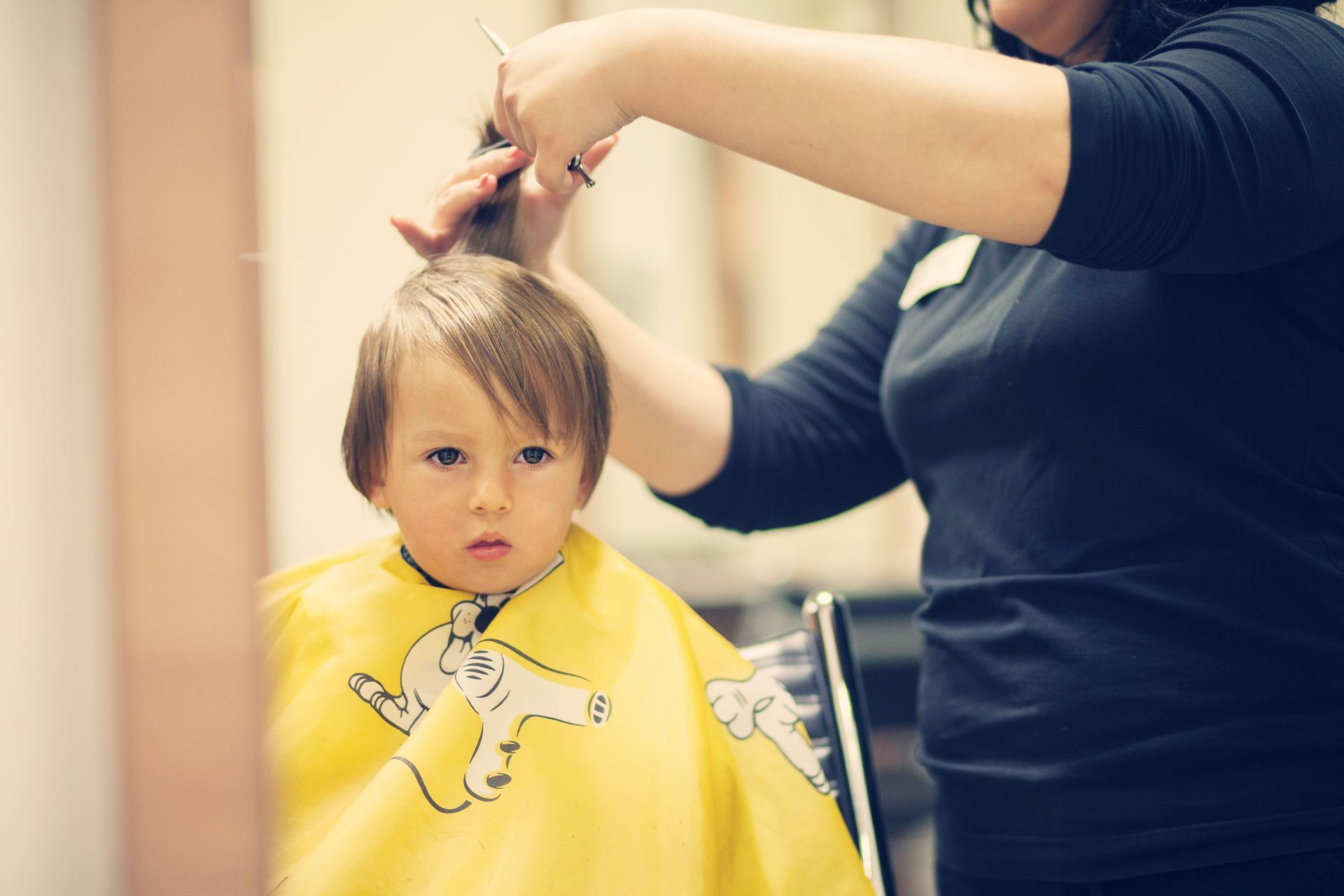 Child getting a haircut in a salon, wearing a yellow cape as a stylist trims their hair.