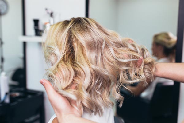 Blonde wavy hair being styled in a salon, viewed from behind with hands holding the curls.