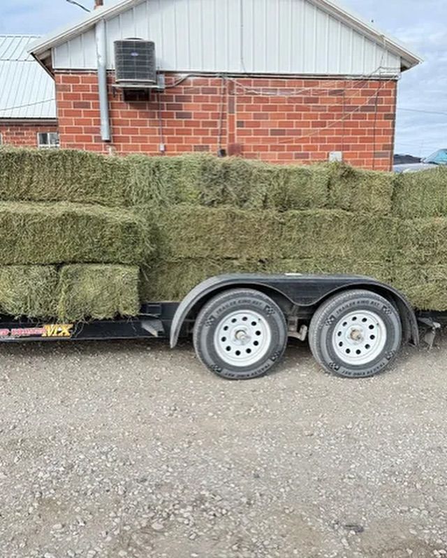 A trailer filled with hay bales is parked in front of a brick building.