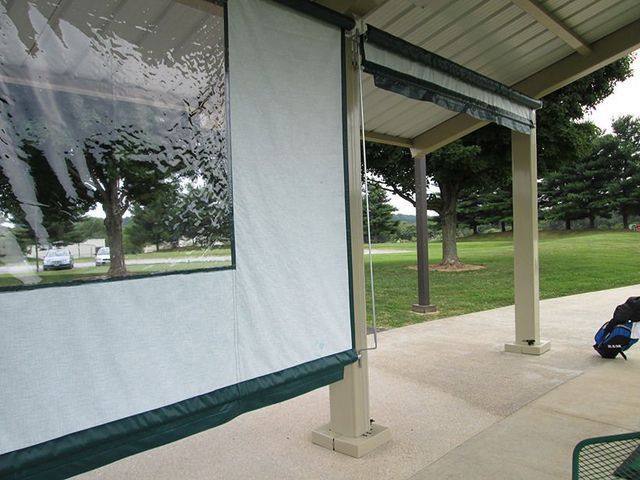 A covered outdoor seating area with white and green roll-down screens. Trees and green space are visible.