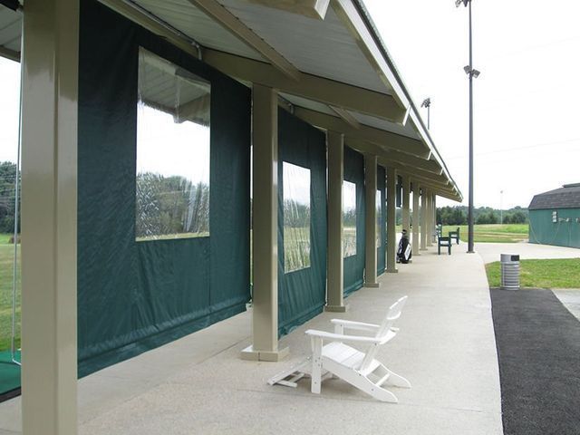 A covered golf driving range with green privacy curtains and white chairs; a green field in the background.