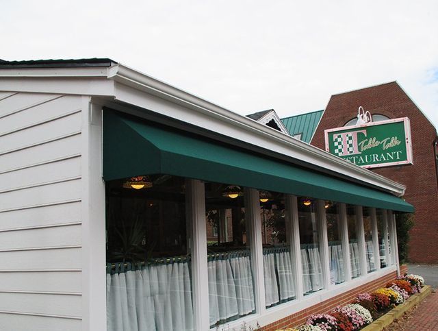 Green awning over a restaurant with a brick building and sign in the background, autumn flowers below.