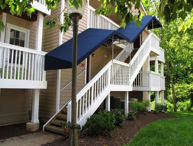 Exterior staircase with blue awning leading to an apartment building entrance. White railing, tan siding, and green grass.