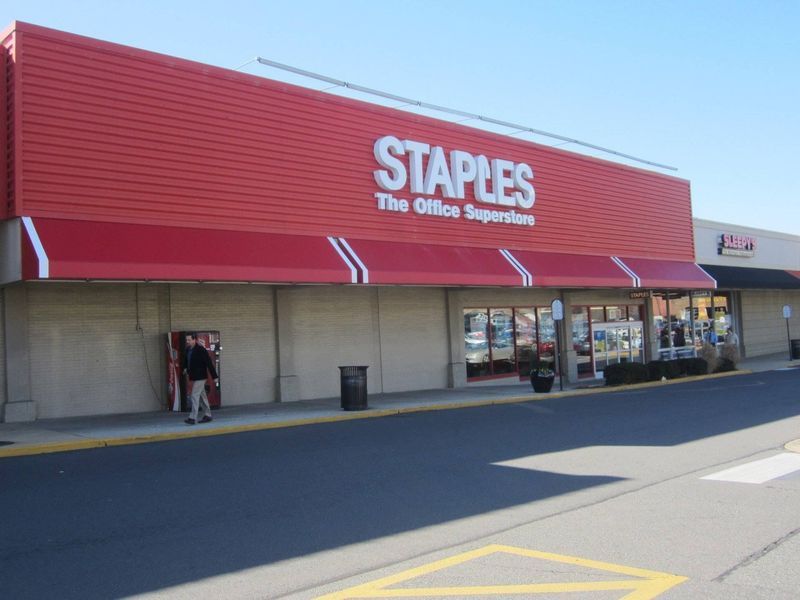 Staples store with red facade, awning, and sign. Person stands outside.