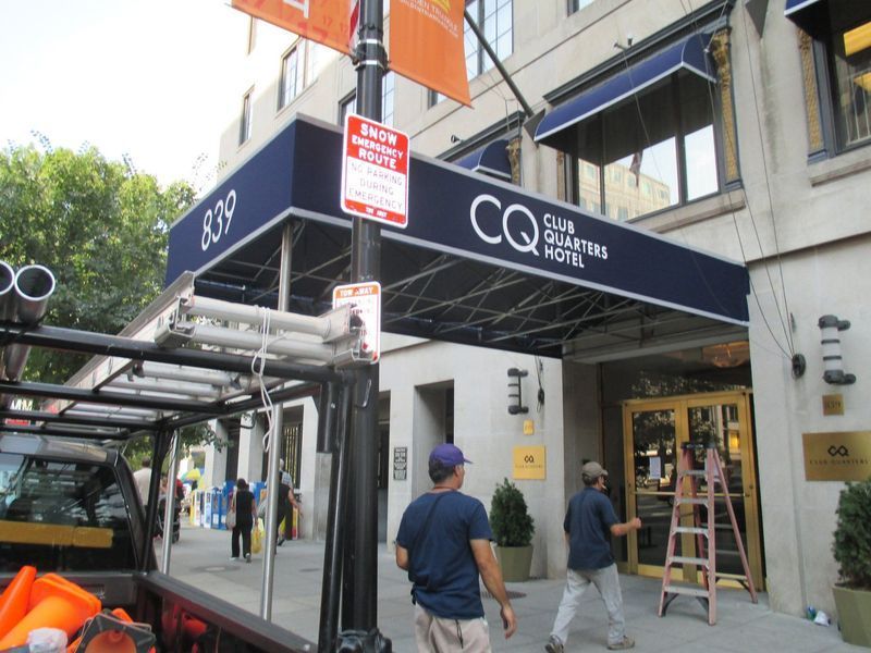 Entrance to CQ Club Quarters Hotel, with workers. Dark blue awning, sign, and storefront.