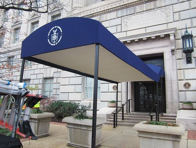 Blue awning over a building entrance with a logo, black metal supports, and stone planters.