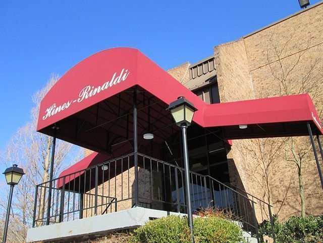 Red awning over the entrance to Rino's Rinaldi restaurant. Black railing, brick building, and clear blue sky.