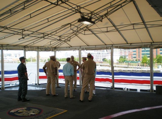 Group of people in naval uniforms and business attire on a deck, possibly a ship, with a water view.