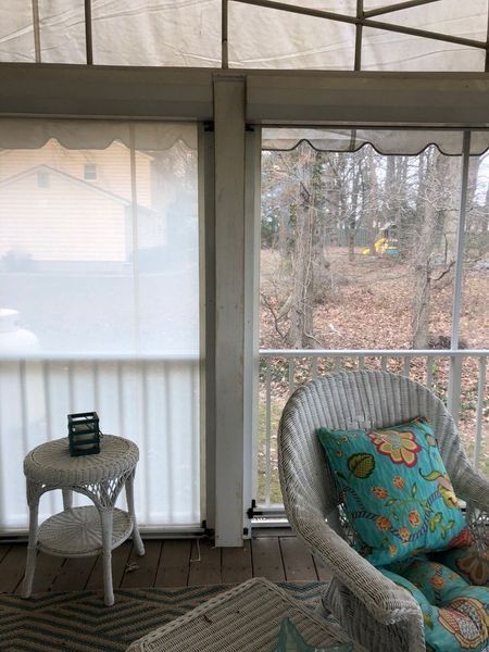 Sunroom interior with white wicker chair and table, white roller shades, and view of trees.