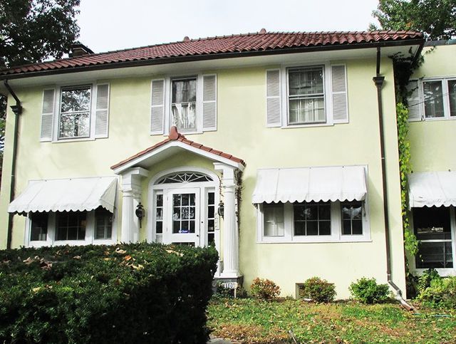 Two-story yellow stucco house with white shutters, awnings, and a red tile roof.