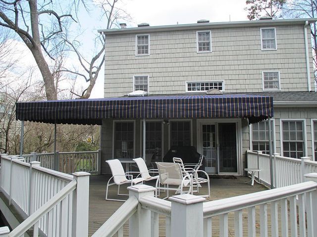 A deck with white railings and several white chairs under a dark awning attached to a two-story house.