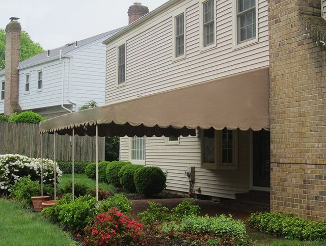 Beige awning covers a house's back porch, supported by white poles, with shrubs and a brick chimney visible.