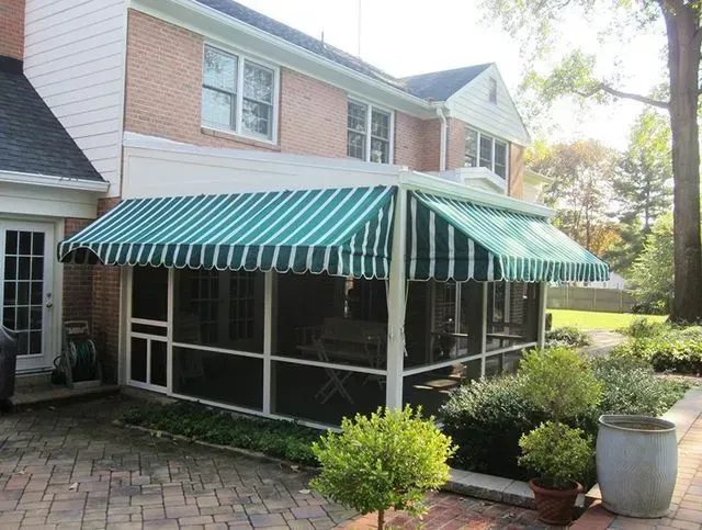 Screened porch with green and white striped awning, attached to a brick house, surrounded by a garden.