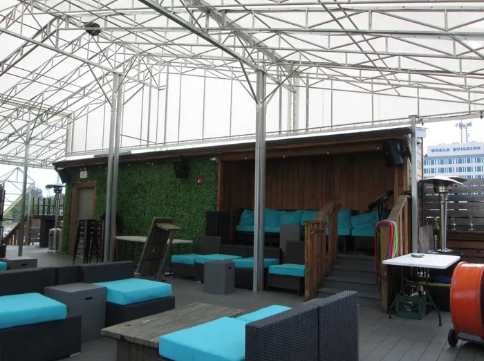 Rooftop patio with turquoise cushions, wooden walls, a white canopy, and a view of a city.