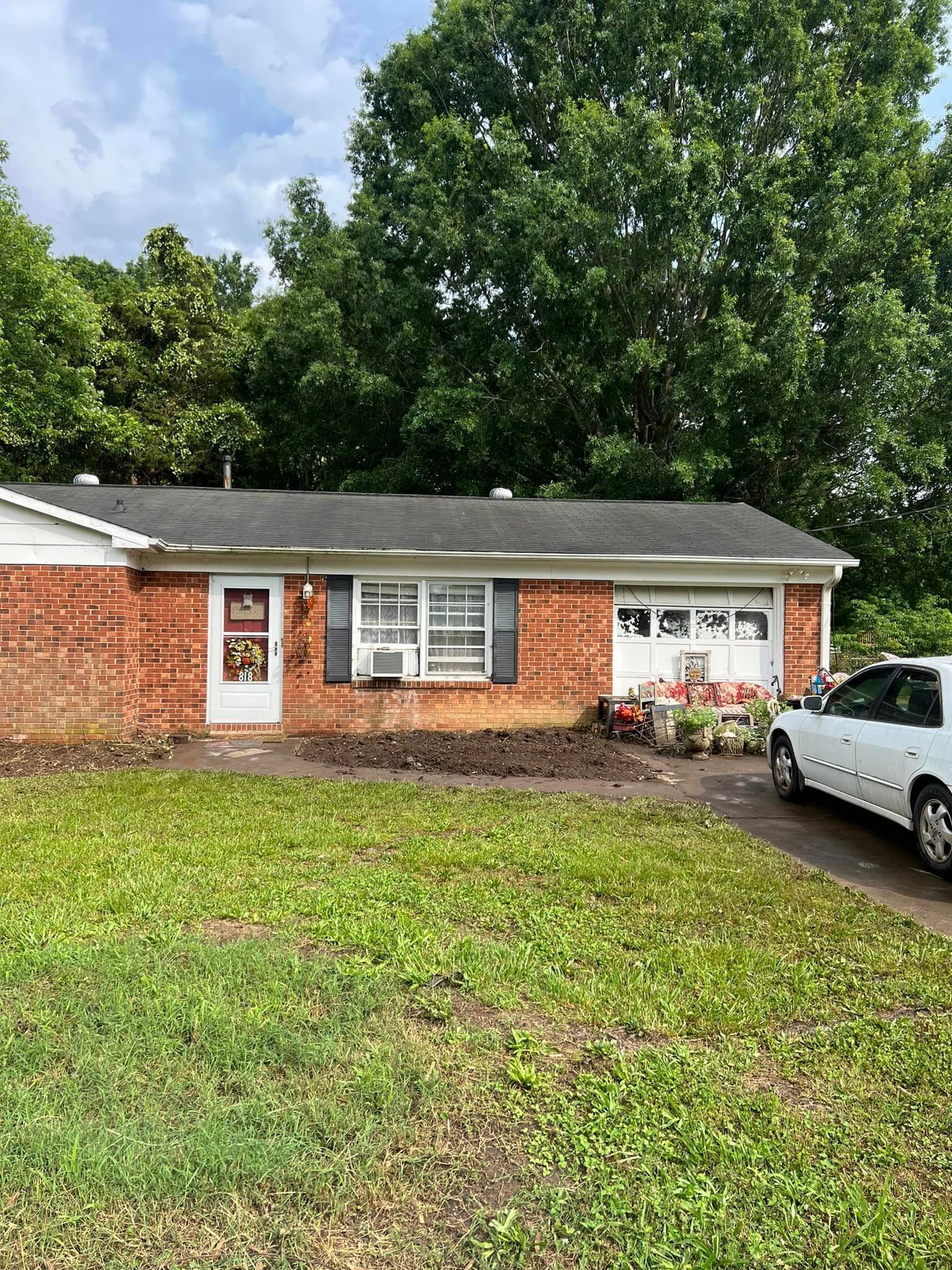 Brick house with a white car parked in the driveway, small front yard with a bare flowerbed, and mature trees in the background.