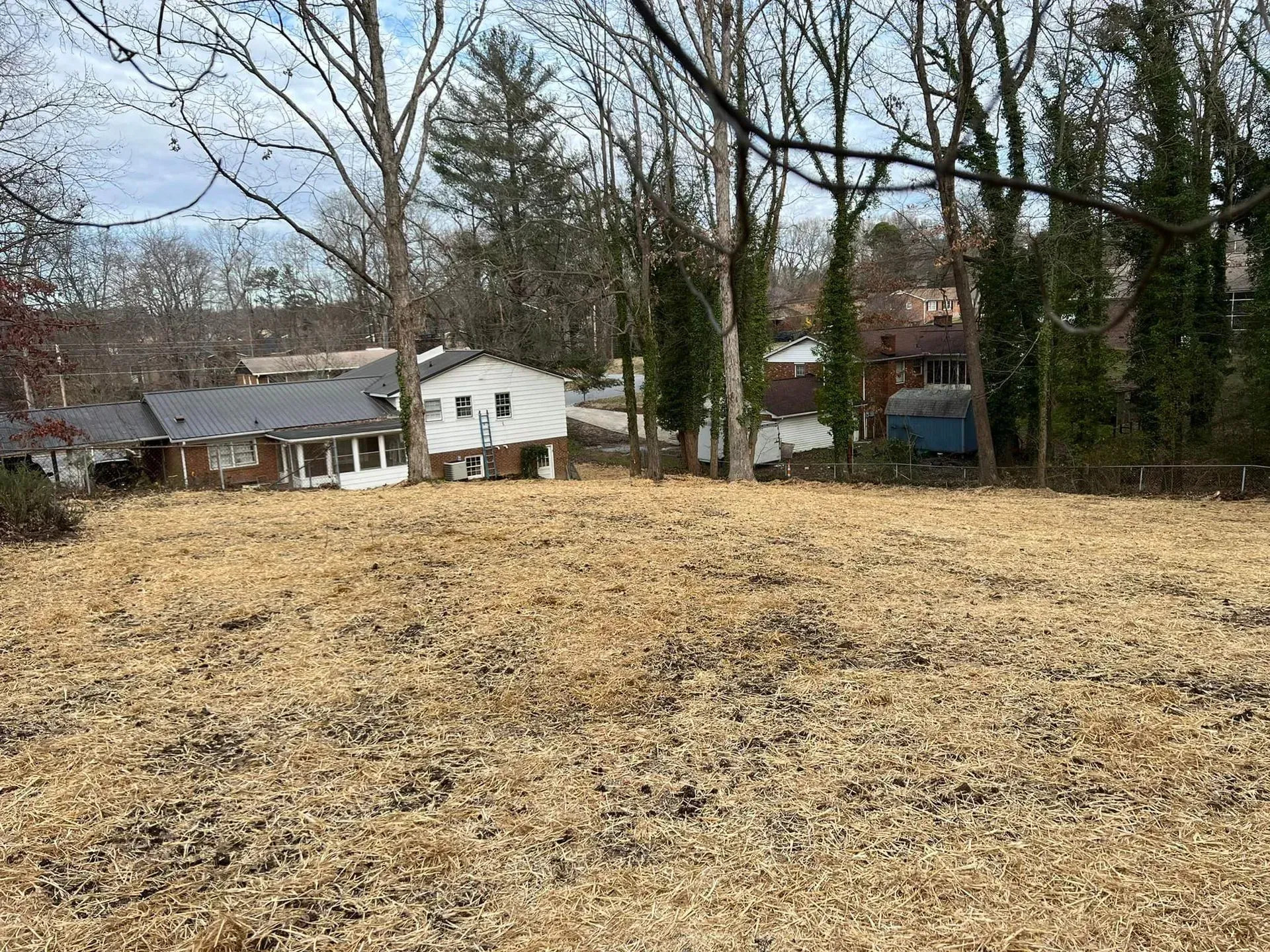 A vacant lot covered in brown dry grass with several houses and trees in the background.