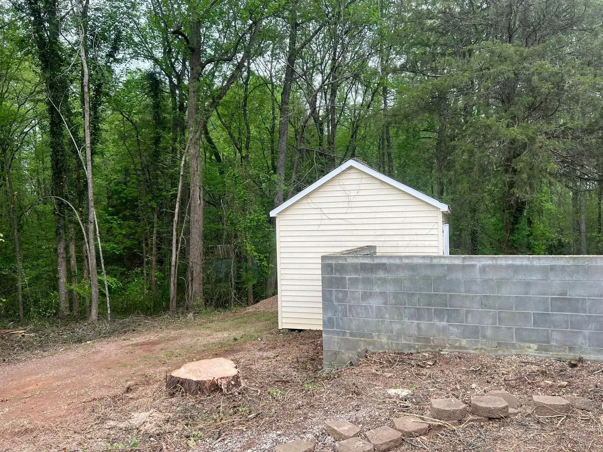 Small light-colored shed beside a gray cinder block wall with trees in the background.