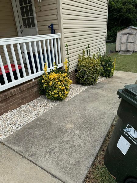 Concrete walkway beside a house with bushes, a white railing, and a trash can.
